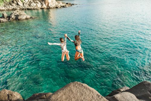 Two people are jumping into clear, blue water from a rocky ledge, holding hands. The sunlit water looks inviting and vibrant.
