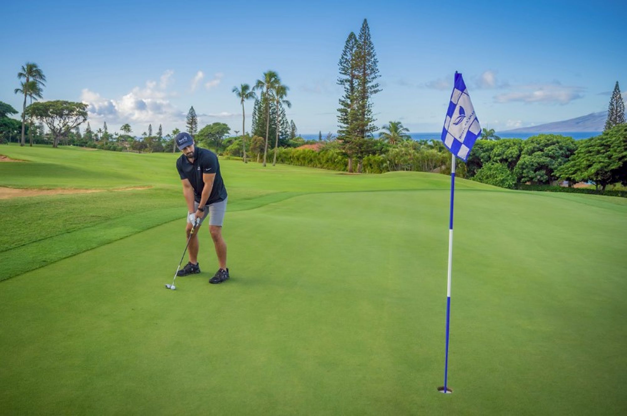 A person is putting on a golf green with a flagstick nearby, surrounded by lush trees and a clear sky in the background.