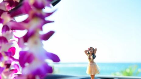 A hula dancer figurine on a car dashboard with an ocean view in the background, and blurred purple flowers in the foreground.