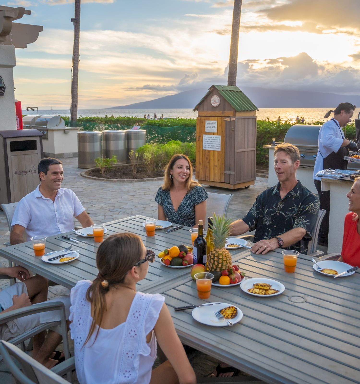 A group of people enjoy a meal outdoors at a table near a beach, with drinks and food on the table, and a person grilling in the background.