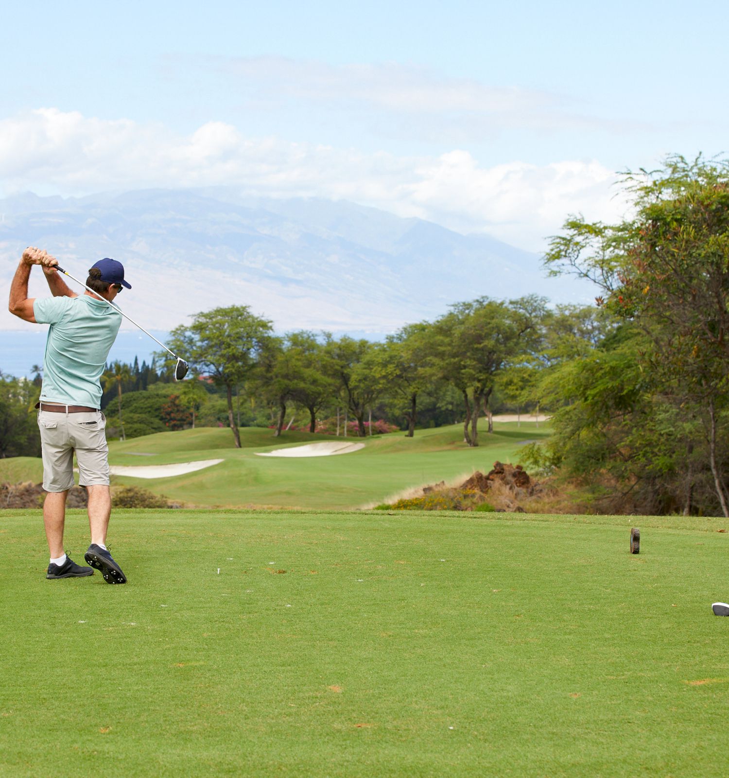 Two people are on a golf course; one is swinging a club, and the other is watching. The background includes trees and distant mountains.