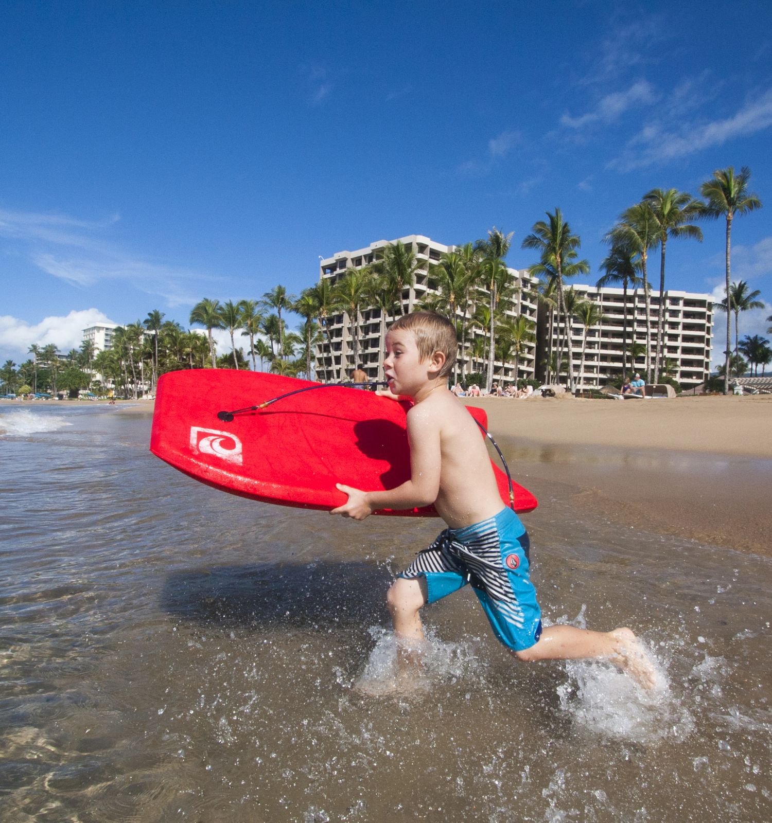 A child runs through the shallow ocean water, holding a red boogie board. Palm trees, beachgoers, and several buildings are visible in the background.