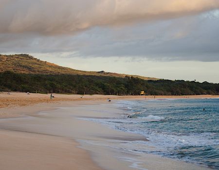 A serene beach with golden sand, calm waves, and a scenic hilly backdrop. People are scattered along the shore under a partly cloudy sky.