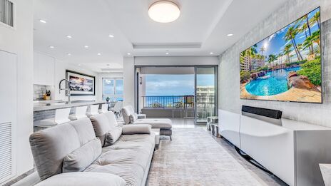 Luxurious coastal living room with light gray sectional, modern white cabinets, panoramic ocean view balcony, and a large wall-mounted TV. End.