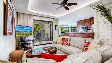 Spacious, modern living room with white sectional sofa, red accent pillows, glass coffee table, ceiling fan, and open layout to a bright kitchen; sliding glass doors lead to a balcony.