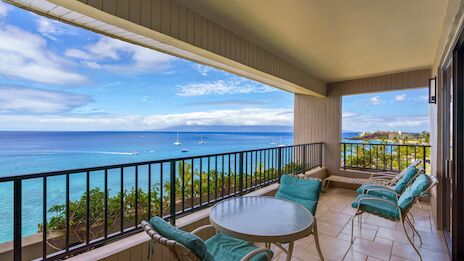 A sunny balcony with turquoise cushioned chairs, a round table, overlooking a blue ocean and distant boats under a clear sky.