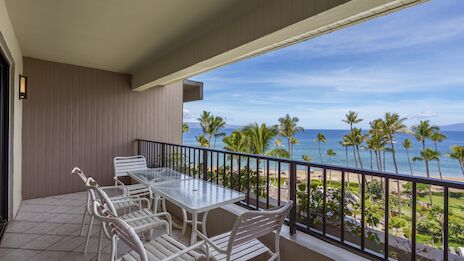 A beachside balcony with a white table and chairs, rail overlooking palm trees and the ocean on a sunny day.