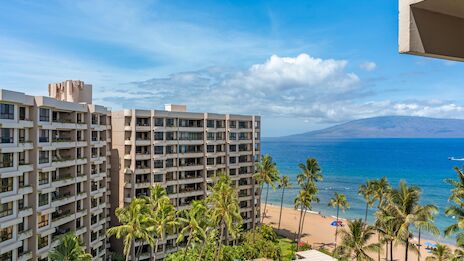 Residential resort buildings by the beach, palm trees, and blue ocean under a sunny sky, with a distant island or hills on the horizon.