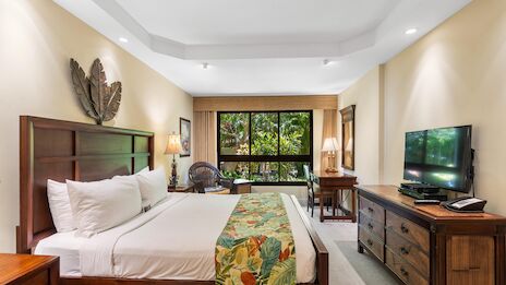 A bright hotel room with a large bed, tropical bed runner, wooden furniture, a desk by a window, a fan, and a flat-screen TV.