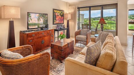 A cozy living room with wicker chairs, a beige sofa, wooden coffee table, a TV on a wooden cabinet, plants, and large windows with a balcony view.