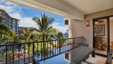 A sunny tropical balcony with a glass table and chairs, palm trees, and a distant building view beyond the railings, under a blue sky.