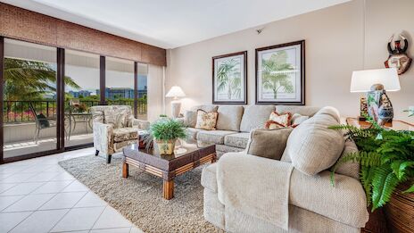 A bright living room with a cozy beige sofa, armchairs, potted plants, and framed art, overlooking a balcony with a scenic view.
