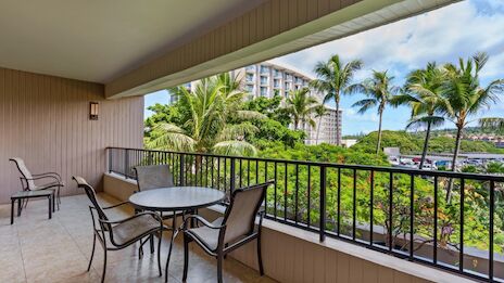 A covered balcony with a round table and four chairs, overlooking palm trees, greenery, and distant buildings; bright, tropical, relaxing.