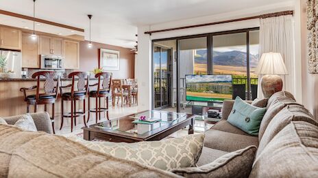 A cozy living room with a sofa, TV, and open kitchen. Large windows showcase mountain views, creating a bright and inviting space.
