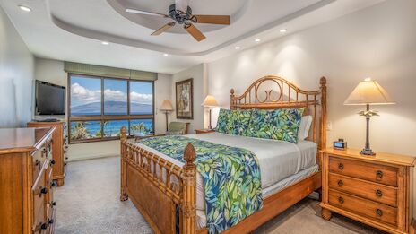A bright tropical hotel room with a wooden bed, matching nightstands, ceiling fan, view of the ocean through large windows, and warm lamps.