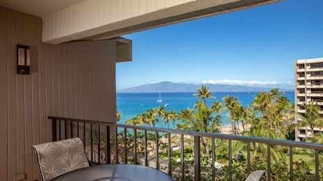 A balcony with a table and chair overlooks a tropical landscape of palm trees and ocean, with a distant island under a clear blue sky.