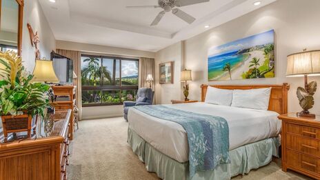 A bright hotel bedroom with a king bed, teal bed scarf, wooden nightstands, ceiling fan, a seating area by a large window, and tropical decor.