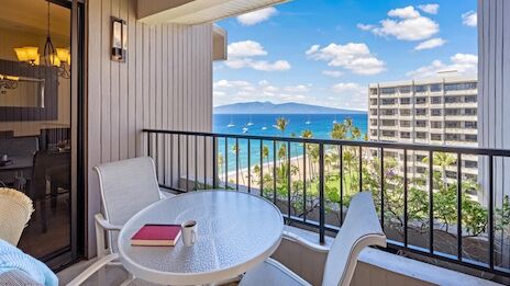 A balcony with a round glass table and four chairs overlooking a blue ocean and blue sky, with buildings and palm trees in view, sunny outdoor scene.