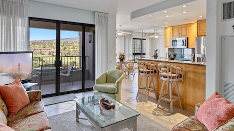 Spacious open-plan living with a floral sofa, glass coffee table, balcony view, and a bright kitchen with wooden cabinets and bar seating, sunlit and airy.