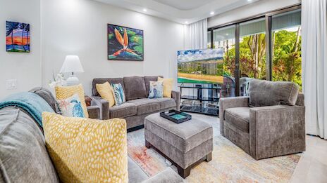 A bright, cozy living room with gray furniture, colorful cushions, artwork, and a balcony view. 140 characters end with a period.