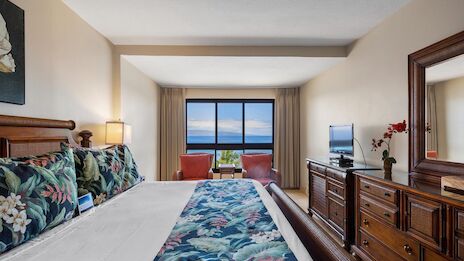 A hotel room with a large bed, tropical floral bedding, nightstands, a dresser with a mirror, and a view of the ocean through a window.