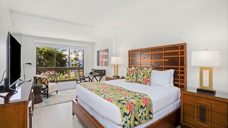 A bright hotel room with a large bed, tropical floral bedding, wooden headboard and furniture, a balcony view, and a desk area across from the TV.