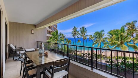 A seaside balcony with a dining table and chairs, palm trees, and a view of the ocean and beach beyond.