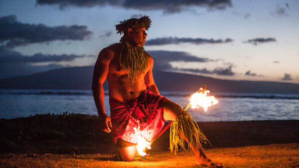 A performer in traditional attire kneels with fire torches, against a scenic backdrop of the ocean and mountains during sunset.