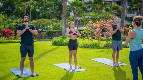 People are practicing yoga outdoors on mats, following an instructor, surrounded by greenery and palm trees.