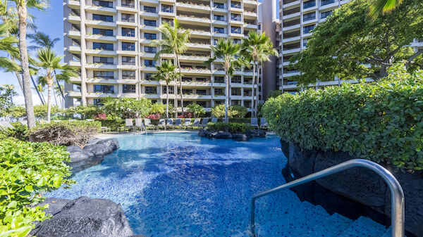 The image features a tropical resort with a clear blue pool surrounded by lush greenery and tall apartment buildings.