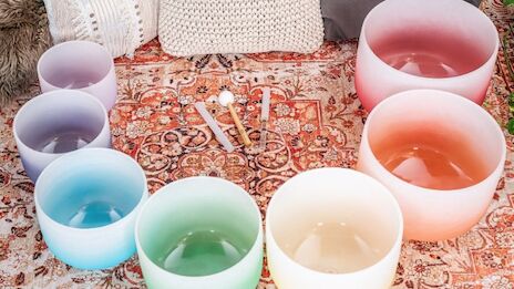 The image features colorful crystal singing bowls arranged on a patterned rug, alongside mallets for playing them.