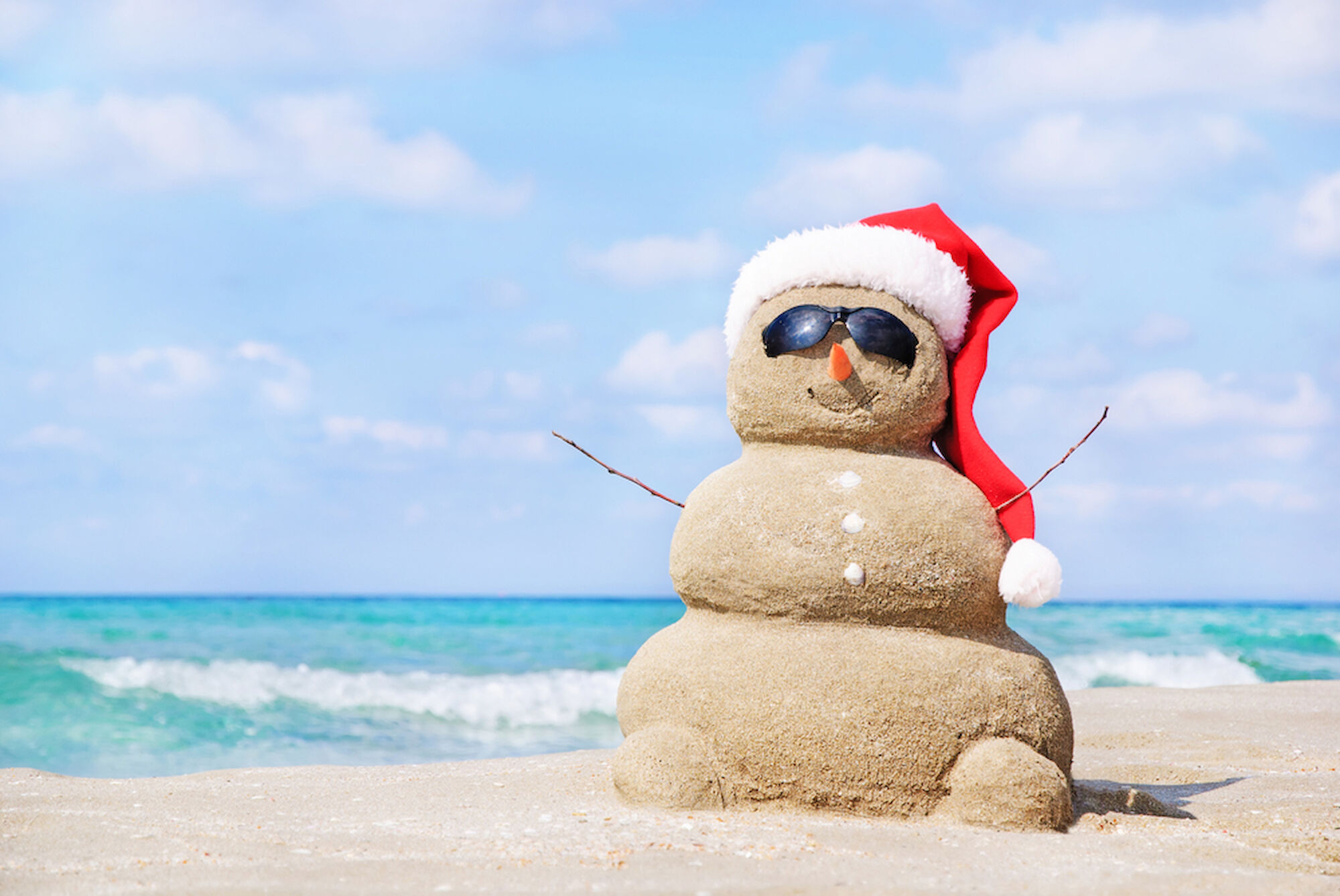 A sand snowman wearing sunglasses and a Santa hat is happily positioned on a sunny beach near the ocean.