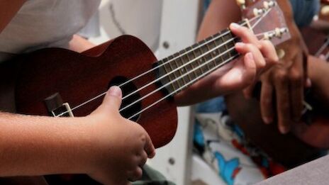 A person playing a small brown ukulele, strumming while others watch, hands on the instrument.