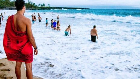 People swim and walk along a tropical beach; a man in a red towel watches the blue waves roll onto the shore.