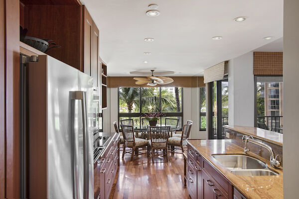 A modern kitchen with stainless steel appliances, wood cabinets, a large island, and a dining area with a view of trees through big windows, bright and airy.