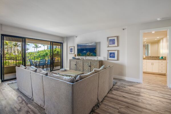Cozy modern living room with a gray sectional, glass sliding door to a balcony, light wood floors, and a hallway leading to a bright kitchen.