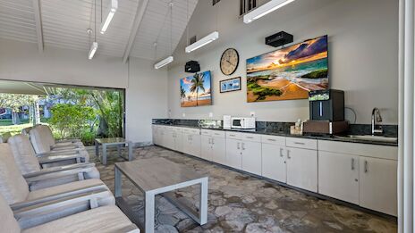 A bright, open lounge/kitchen area with white cabinets, a long counter, coffee maker, mounted artwork, a glass coffee table, and white chairs along a tiled floor.