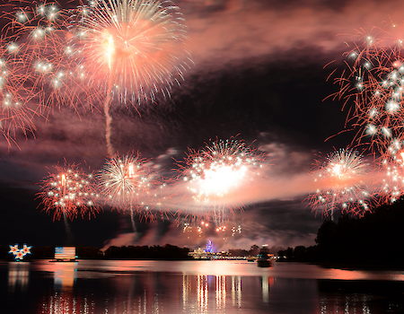 Fireworks explode over a dark lake at night, reflecting colorful bursts and smoke across the water.