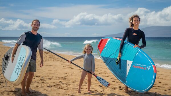 A family stands on a sunny beach with three people holding surfboards: a man and a woman with a child between them, all ready to surf.