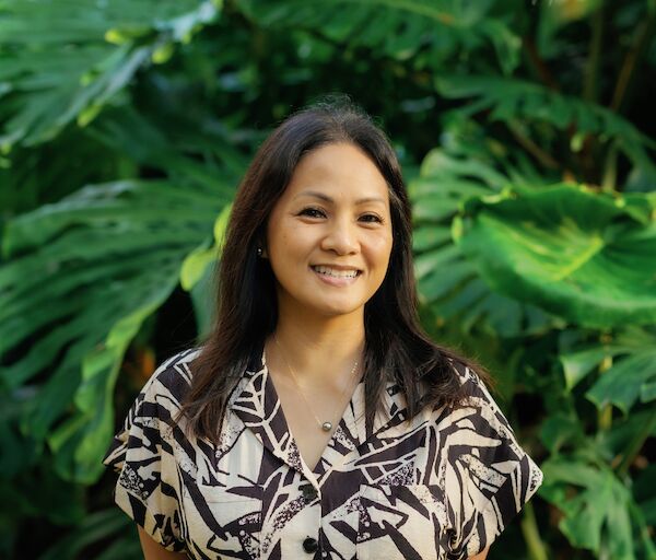 A smiling woman stands outdoors in a patterned dress, with lush green tropical plants in the background. }