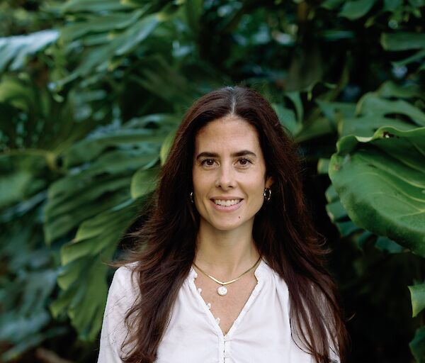 A woman stands outdoors among large green leaves, wearing a white blouse and dark pants, smiling softly with layered jewelry.