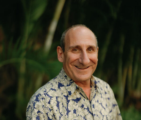 A smiling middle-aged man in a light, tropical-patterned shirt stands outdoors among lush greenery.