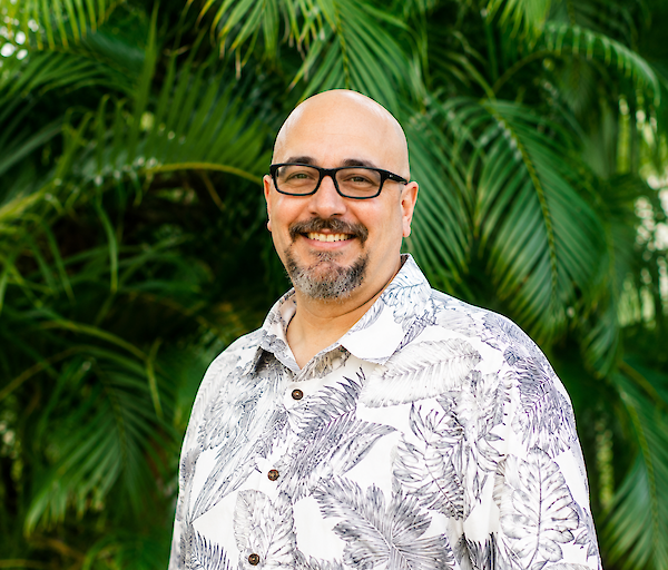 A smiling man with glasses and a patterned short-sleeve shirt stands outdoors, lush green tropical plants in the background.