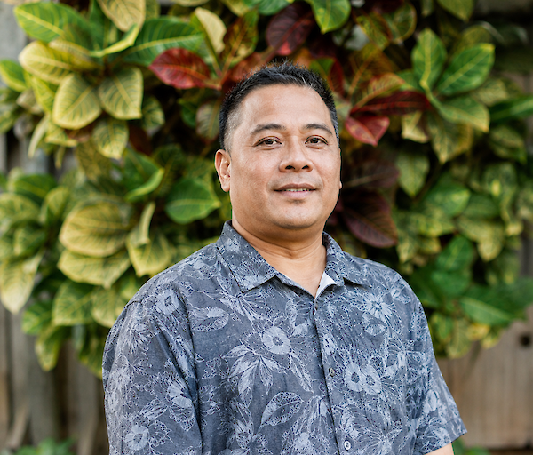 A man in a blue patterned short-sleeve shirt stands outdoors in front of a green and yellow leafy background.