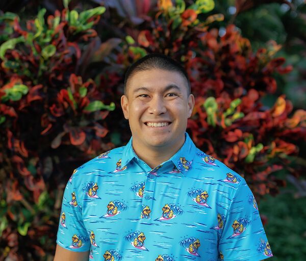 A smiling man stands outdoors in a bright blue short-sleeve shirt with playful cartoon patterns, hands in pockets, tropical plants behind him.