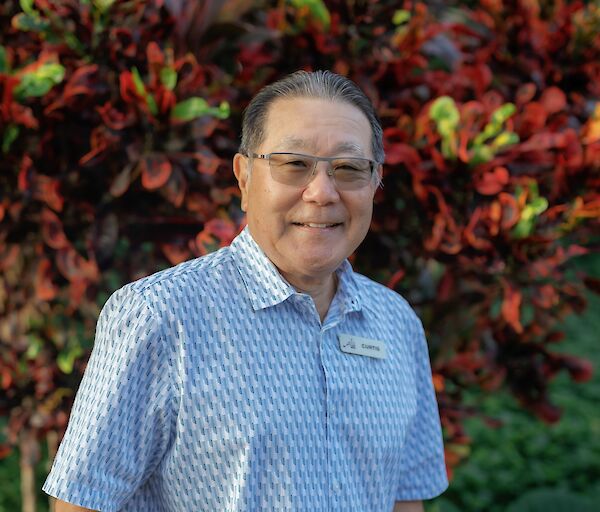A smiling man in a light blue short-sleeve patterned shirt stands outdoors with tropical plants in the background, hands in pockets.