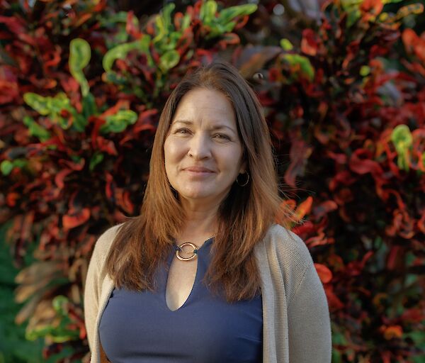 A woman with brown hair stands outdoors, wearing a navy top and light cardigan, against colorful red and green foliage, smiling softly.