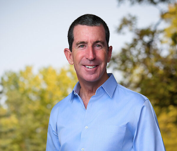 A smiling man in a light blue dress shirt and dark trousers stands outdoors in a park, with trees and fall foliage in the background.