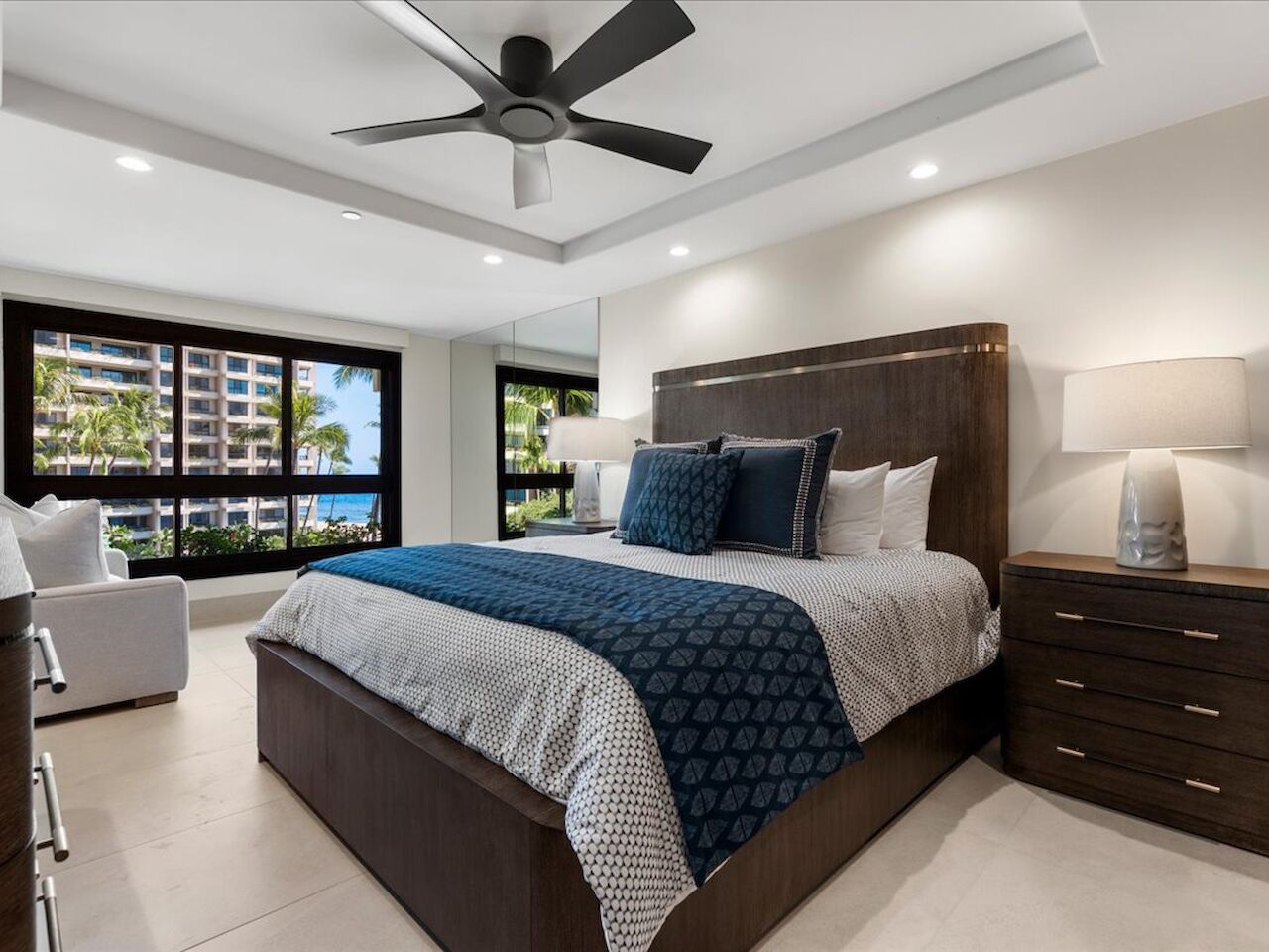 A modern bedroom with a dark wood bed, blue and white bedding, matching nightstands, a ceiling fan, recessed lights, and a large window overlooking a city view.