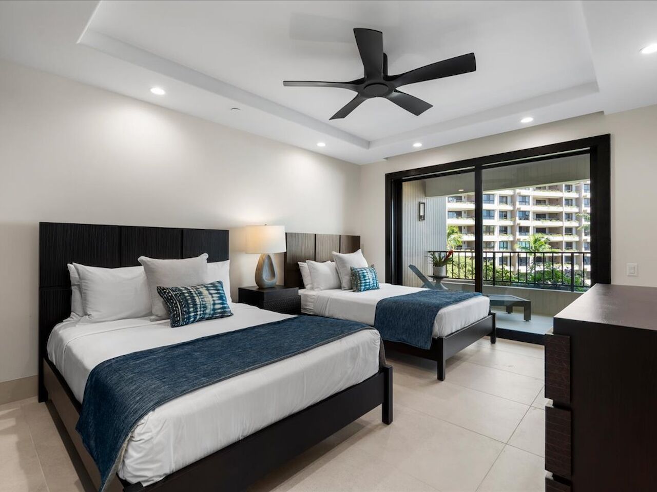 A bright hotel room with two queen beds, blue runners and pillows, a dark wood dresser, a ceiling fan, and a balcony view of nearby buildings.